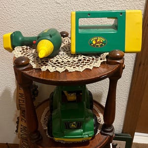 May include: A wooden three-tiered stand with a green toy tractor, a green toy drill, and a green and yellow toy flashlight. The flashlight has the John Deere Kids logo on it.
