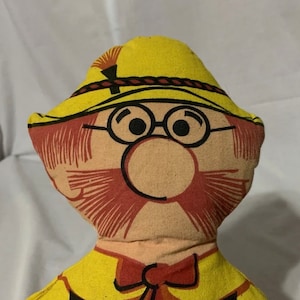 May include: A vintage fabric doll with a cartoonish face. The doll has a yellow hat, glasses, and a red mustache and bow tie. The doll's face is printed on a tan fabric.