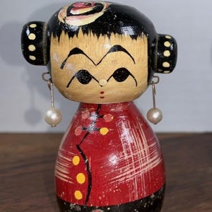 May include: A vintage wooden doll with a painted face, black hair, and a red dress with yellow accents. The doll has pearl earrings and is a collectible item.
