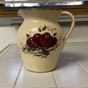 以下が含まれることがあります： 緑色の縁取りと取っ手のあるクリーム色のセラミックピッチャー。ピッチャーには、赤いリンゴと緑の葉の手描きデザインが施されています。