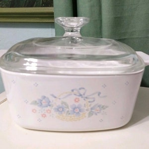 Puede incluir: Plato para hornear blanco con un diseño floral que presenta flores azules, una cesta y cintas rosas y azules. El plato tiene una tapa de vidrio transparente con un pomo.