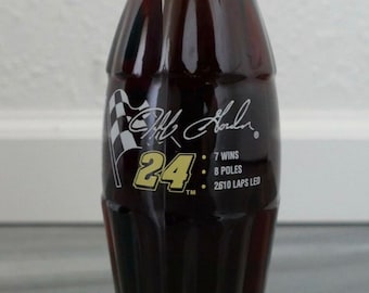May include: A glass Coca-Cola bottle with a red cap and a black label featuring a white checkered flag, the number 24 in gold, and the signature of Jeff Gordon. The label also includes the text "7 Wins", "8 Poles", and "2610 Laps Led".