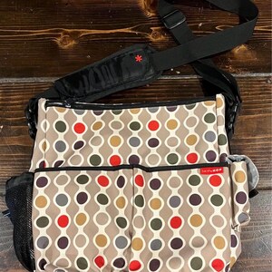 Puede incluir: Un bolso cambiador beige con un patr&oacute;n repetitivo de c&iacute;rculos coloridos en rojo, marr&oacute;n, gris y amarillo. El bolso tiene una correa para el hombro negra y m&uacute;ltiples bolsillos, incluido un bolsillo lateral de malla. La marca "SKIP HOP" es visible.