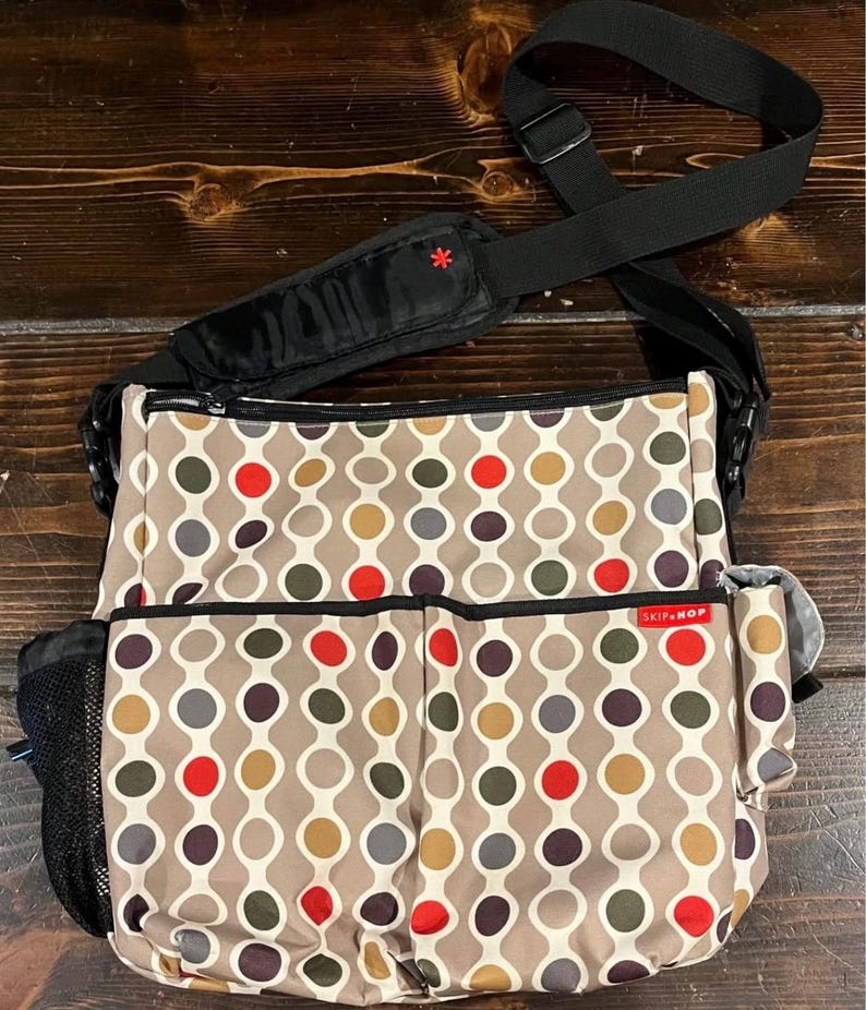 Puede incluir: Un bolso cambiador beige con un patr&oacute;n repetitivo de c&iacute;rculos coloridos en rojo, marr&oacute;n, gris y amarillo. El bolso tiene una correa para el hombro negra y m&uacute;ltiples bolsillos, incluido un bolsillo lateral de malla. La marca "SKIP HOP" es visible.