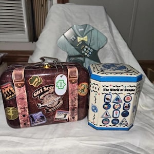 Puede incluir: Una colección de recuerdos vintage de las Girl Scouts, que incluye una lata en forma de maleta con pegatinas de viaje, una lata en forma de uniforme y una lata hexagonal con el texto "The World of People."
