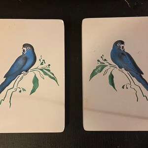 May include: Two white ceramic tiles with a blue bird perched on a green branch. The bird has a black beak and black eyes.