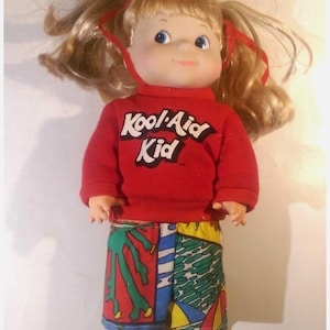 May include: A red and white doll wearing a red sweatshirt with the words "Kool-Aid Kid" on the front and colorful patterned pants.
