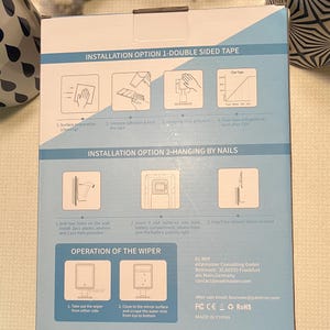 May include: A white box with blue and black text detailing the installation and operation of a bathroom mirror with a built-in wiper. The box includes instructions for installing the mirror using double-sided tape or nails. The box also includes instructions for using the wiper to remove water mist from the mirror.