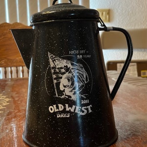 May include: Black enamel coffee pot with a white graphic of a cowboy holding an American flag. The graphic includes the text "Hats Off - 20 Years" and "Old West Days 2011".