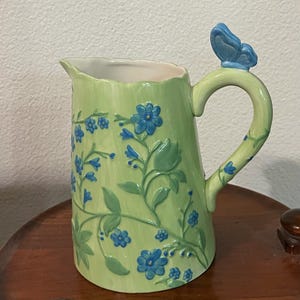 May include: A green ceramic pitcher with a butterfly handle and blue floral design. The pitcher has a textured surface and a wide mouth.