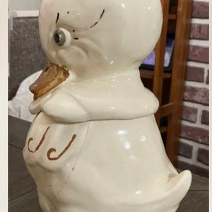May include: A white ceramic cookie jar shaped like a duck with brown painted details. The duck has a large beak and a small eye. The cookie jar is in the shape of a duck sitting down.