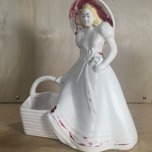 May include: White ceramic planter shaped like a woman wearing a red and white hat and a white dress. The woman is holding a basket in her hand. The planter has a distressed look with some of the paint chipped off.