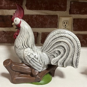 May include: A white ceramic rooster figurine with a red comb and wattle, perched on a brown wooden base with green grass. The rooster's tail feathers are curled and detailed.