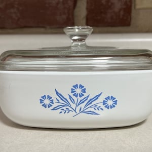 Puede incluir: Plato para hornear blanco con un diseño floral azul y una tapa de vidrio transparente. El plato tiene una forma redondeada y dos asas.