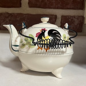 May include: White ceramic teapot with a rooster design and a black metal handle. The teapot has three legs and a spring-loaded lid.