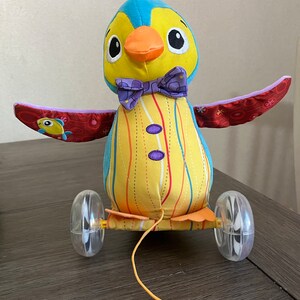 May include: A yellow and blue penguin pull toy with red wings and a purple bow tie. The penguin has black eyes and an orange beak. The toy has clear plastic wheels and a yellow string.