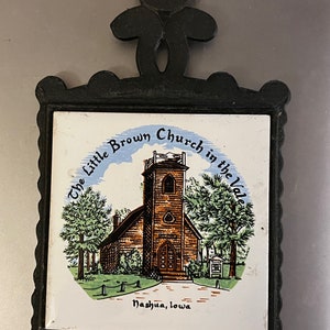May include: A black cast iron hot pad with a white ceramic tile insert. The tile features a colorful illustration of a church with the text "The Little Brown Church in the Vale Nashua, Iowa".