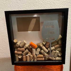 Könnte beinhalten: Eine schwarze Schattenbox mit einer Glasfront, gefüllt mit Weinkorken. Die Box hat einen weißen, mattierten Bilderrahmen und einen silbernen Vinyl-Aufkleber eines Weinglases mit dem Text "Gute Freunde, gute Zeiten, guter Wein".