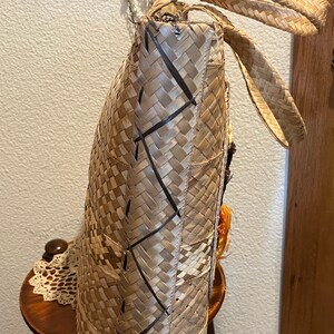 K&ouml;nnte beinhalten: Eine geflochtene Strohtasche mit braunen und schwarzen Akzenten. Die Tasche hat eine gro&szlig;e, rechteckige Form und zwei lange Henkel. Die Tasche ist aus nat&uuml;rlichen Materialien gefertigt und hat einen rustikalen, handgefertigten Look.
