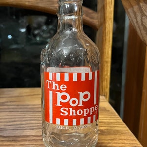 Könnte beinhalten: Eine durchsichtige Glasflasche mit einem roten und weißen Etikett, auf dem "The Pop Shoppe" und "10.14 FL. OZ./300 ml" steht.