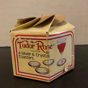 May include: A box of four silver and crystal coasters from the Tudor Rose Collection. The box is yellow with red and purple stripes and features a glass of red wine.