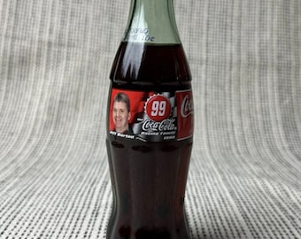 May include: A glass Coca-Cola bottle with a red cap featuring a picture of Jeff Burton, the Coca-Cola Racing Family, and the number 99. The bottle is filled with dark brown liquid.