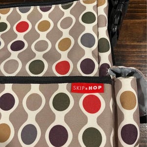 Puede incluir: Un bolso para pa&ntilde;ales Skip Hop con un patr&oacute;n repetitivo de c&iacute;rculos de colores en rojo, verde, morado y marr&oacute;n sobre un fondo gris. El bolso tiene una correa negra y una etiqueta roja con el logotipo de Skip Hop.
