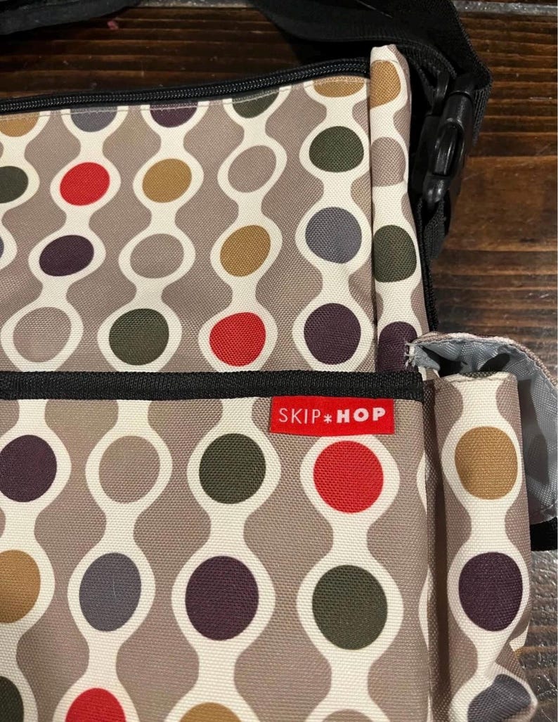 Puede incluir: Un bolso para pa&ntilde;ales Skip Hop con un patr&oacute;n repetitivo de c&iacute;rculos de colores en rojo, verde, morado y marr&oacute;n sobre un fondo gris. El bolso tiene una correa negra y una etiqueta roja con el logotipo de Skip Hop.
