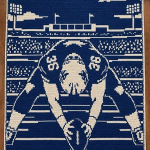 May include: A rectangular crochet blanket featuring a two-tone blue and cream design. The graphic depicts a football player in a jersey with the number 36, positioned at the line of scrimmage on a field with stadium lights and clouds above.