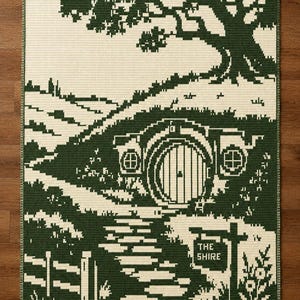 Könnte beinhalten: Eine rechteckige Häkeldecke mit einem pixeligen Landschaftsmotiv in Dunkelgrün und Creme. Die Szene zeigt eine runde Tür in einem grasbewachsenen Hügel unter einem großen Baum sowie einen Steinpfad, der zu einem Schild mit der Aufschrift THE SHIRE führt.