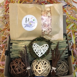 May include: A brown box filled with a variety of small animal enrichment items, including a bag of dried fruit, a heart-shaped woven decoration, a bundle of sticks, a woven ball, a brown ball, and a bed of hay. The box is labeled "Nibbles Store".
