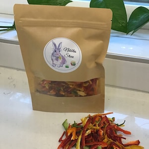 May include: A brown paper bag with a clear window, filled with colorful dried vegetables. A sticker on the bag features a purple bunny and the text "Nibbles Store." A pile of the dried vegetables sits in front of the bag.