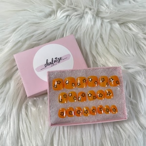 May include: A set of orange press-on nails with black star designs, displayed in a pink box. The box has a white circle with the text "chalaize nails" in a cursive font. The nails are arranged on a white, fluffy surface.