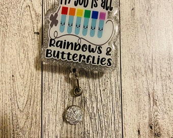 May include: A silver glitter badge holder with a clear plastic charm featuring the text "My job is all rainbows & butterflies" and a row of colourful test tubes with smiley faces.