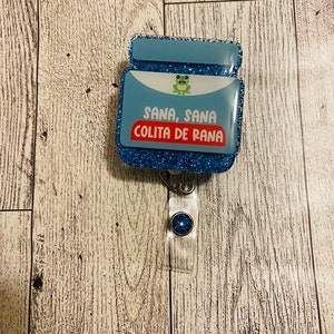 May include: A blue glitter badge reel featuring a rectangular acrylic topper with a light blue background, a small green frog illustration, and the text "SANA, SANA COLITA DE RANA" printed in white and red letters on a clear plastic strap.
