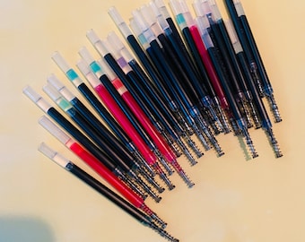 May include: A collection of black and clear pen refills with red, blue, and green ink. The refills are arranged in a fan shape on a light yellow surface.
