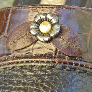 May include: A dark brown shoe with a crocodile-embossed texture featuring a decorative silver-toned metal flower ornament with a gold-toned center, accented by two brown leather leaf-shaped appliqués stitched onto the upper section.