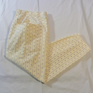 May include: Cream-colored pants with a vertical striped pattern of small, dark blue circles and lines. The pants have a pocket on the back and are folded. The fabric appears to be lightweight.