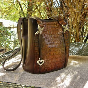 May include: A brown leather handbag with a crocodile-skin texture on the front. The bag has a shoulder strap and a top handle. Silver-colored metal accents and a heart-shaped charm add detail to the design. The bag is sitting on a light brown surface.