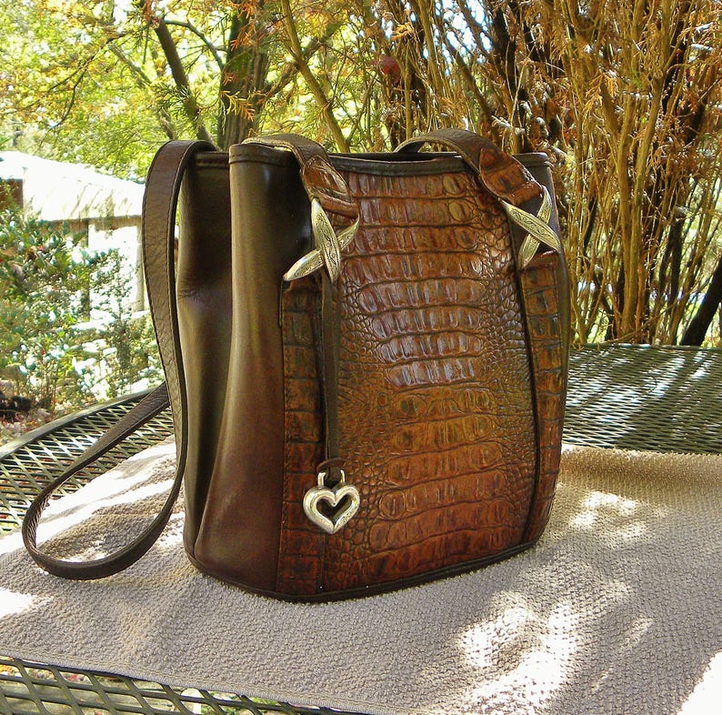 May include: A brown leather handbag with a crocodile-skin texture on the front. The bag has a shoulder strap and a top handle. Silver-colored metal accents and a heart-shaped charm add detail to the design. The bag is sitting on a light brown surface.