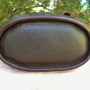 May include: Close-up of the bottom of a dark brown leather handbag. The oval base has a textured surface and is framed by a darker brown, smooth leather trim. The bag appears to be well-crafted and durable.