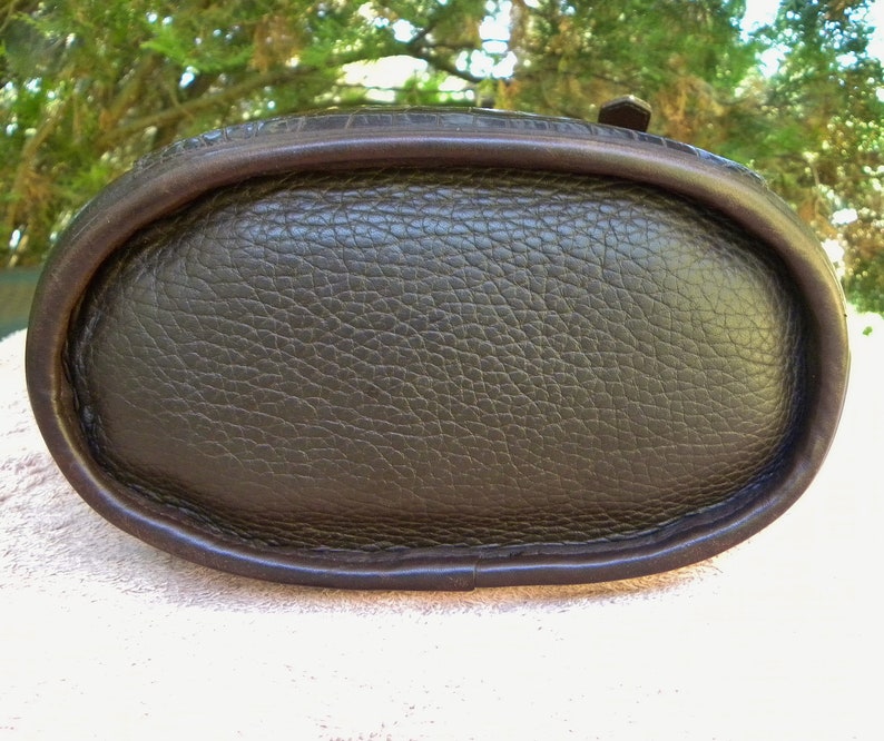 May include: Close-up of the bottom of a dark brown leather handbag. The oval base has a textured surface and is framed by a darker brown, smooth leather trim. The bag appears to be well-crafted and durable.
