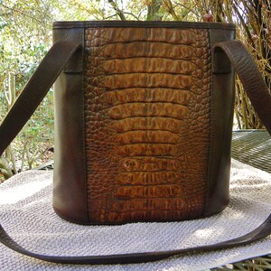 May include: A brown leather handbag with a crocodile-skin texture on the front panel. The bag has a rectangular shape with a long, thin strap. The sides and strap are a darker brown than the front panel.