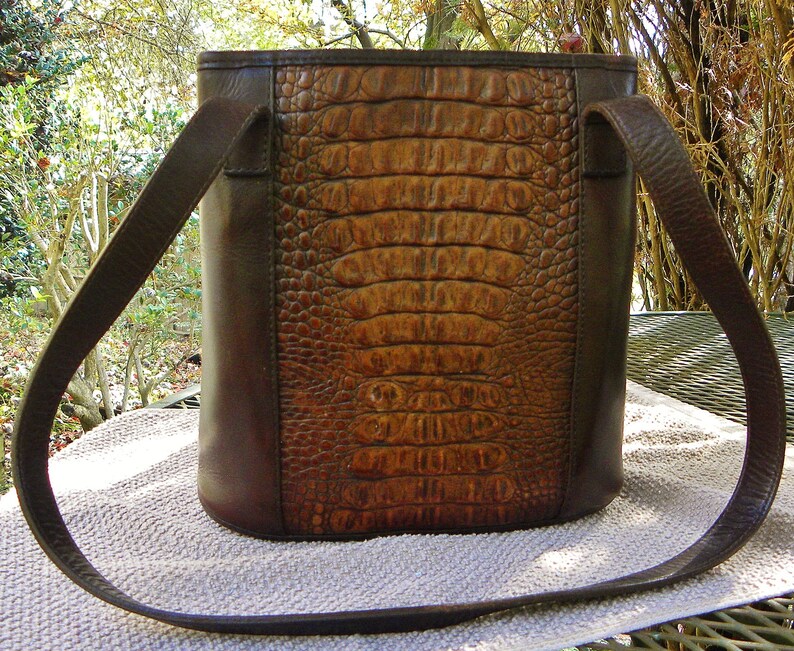May include: A brown leather handbag with a crocodile-skin texture on the front panel. The bag has a rectangular shape with a long, thin strap. The sides and strap are a darker brown than the front panel.