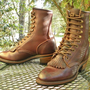 May include: A pair of brown leather lace-up boots featuring a mid-calf height, brass-toned eyelets, and a decorative fringe panel over the vamp. The boots have a stacked wooden heel, a rounded toe, and brown laces threaded through the eyelets.
