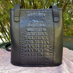 May include: Black tote bag with a faux crocodile skin center panel and smooth black sides. The bag has two top handles and a shoulder strap. The bag is sitting on a pink towel.