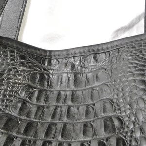 May include: Black leather tote bag with a crocodile-textured center panel. The bag features silver-toned decorative accents and sturdy black straps. The bag's design is both stylish and functional.
