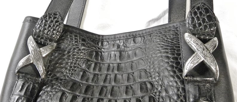 May include: Black leather tote bag with a crocodile-textured center panel. The bag features silver-toned decorative accents and sturdy black straps. The bag's design is both stylish and functional.