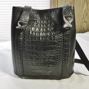 May include: Black bucket bag with a crocodile-textured center panel and smooth side panels. The bag features silver-tone hardware on the straps and a long, adjustable strap for shoulder or crossbody wear.