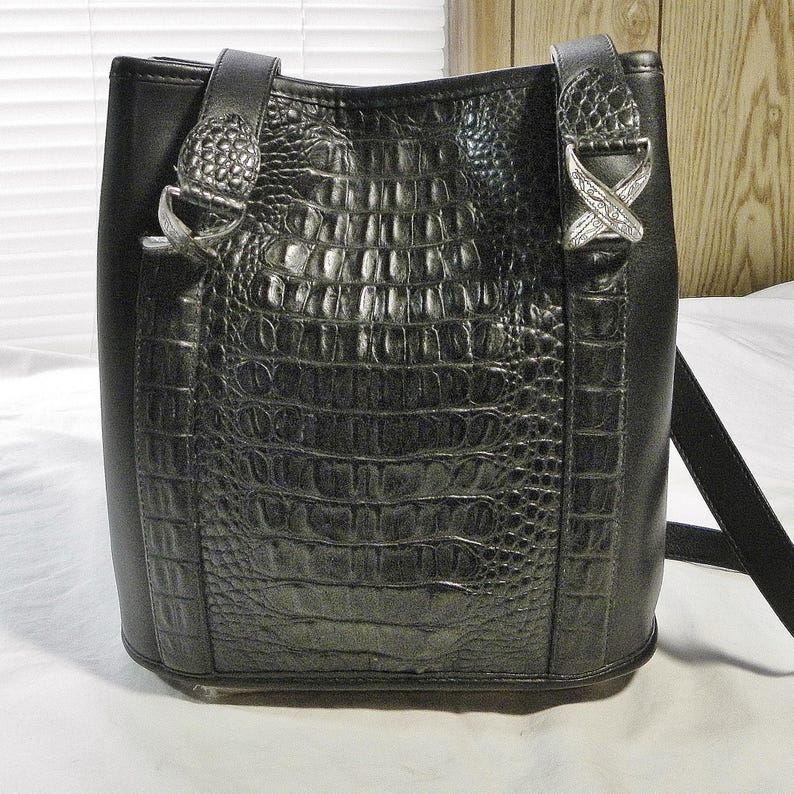 May include: Black bucket bag with a crocodile-textured center panel and smooth side panels. The bag features silver-tone hardware on the straps and a long, adjustable strap for shoulder or crossbody wear.